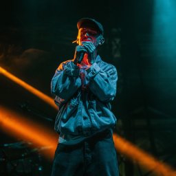 ENEN STUDIO - Młody mężczyzna w dżinsowej kurtce i czapce rapera, śpiewa do mikrofonu na scenie oświetlonej pomarańczowym i turkusowym światłem.