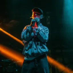 Młody mężczyzna w dżinsowej kurtce i czapce rapera, śpiewa do mikrofonu na scenie oświetlonej pomarańczowym i turkusowym światłem.