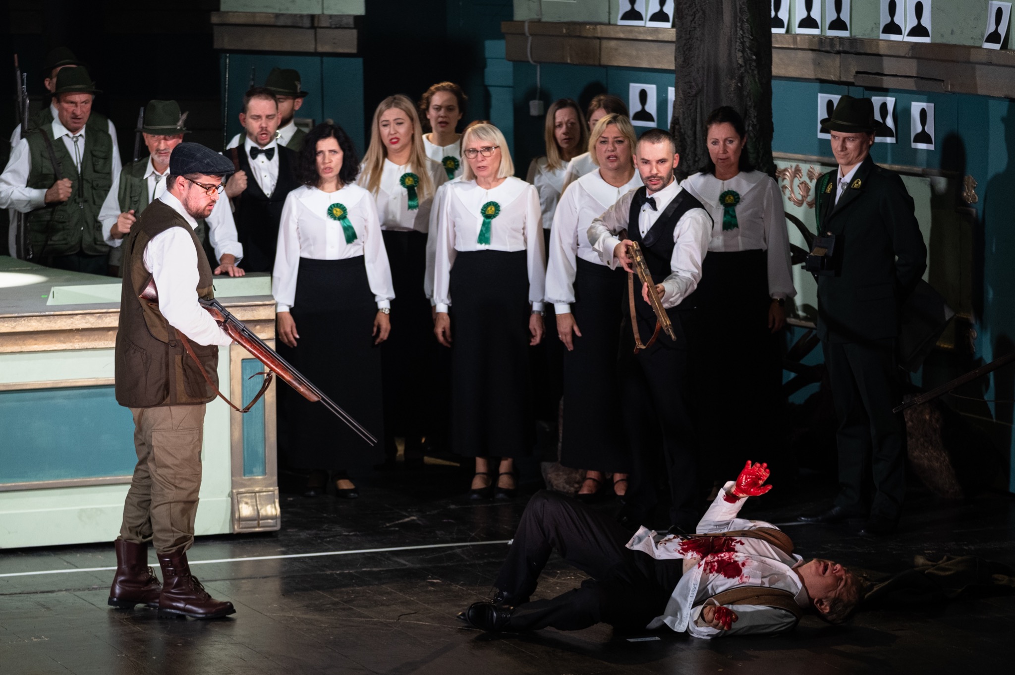 Scena z przedstawienia teatralnego: mężczyzna w brązowej kamizelce trzyma strzelbę, obok leży postrzelony mężczyzna z zakrwawioną ręką, w tle chór w białych bluzkach z zielonymi rozetami...