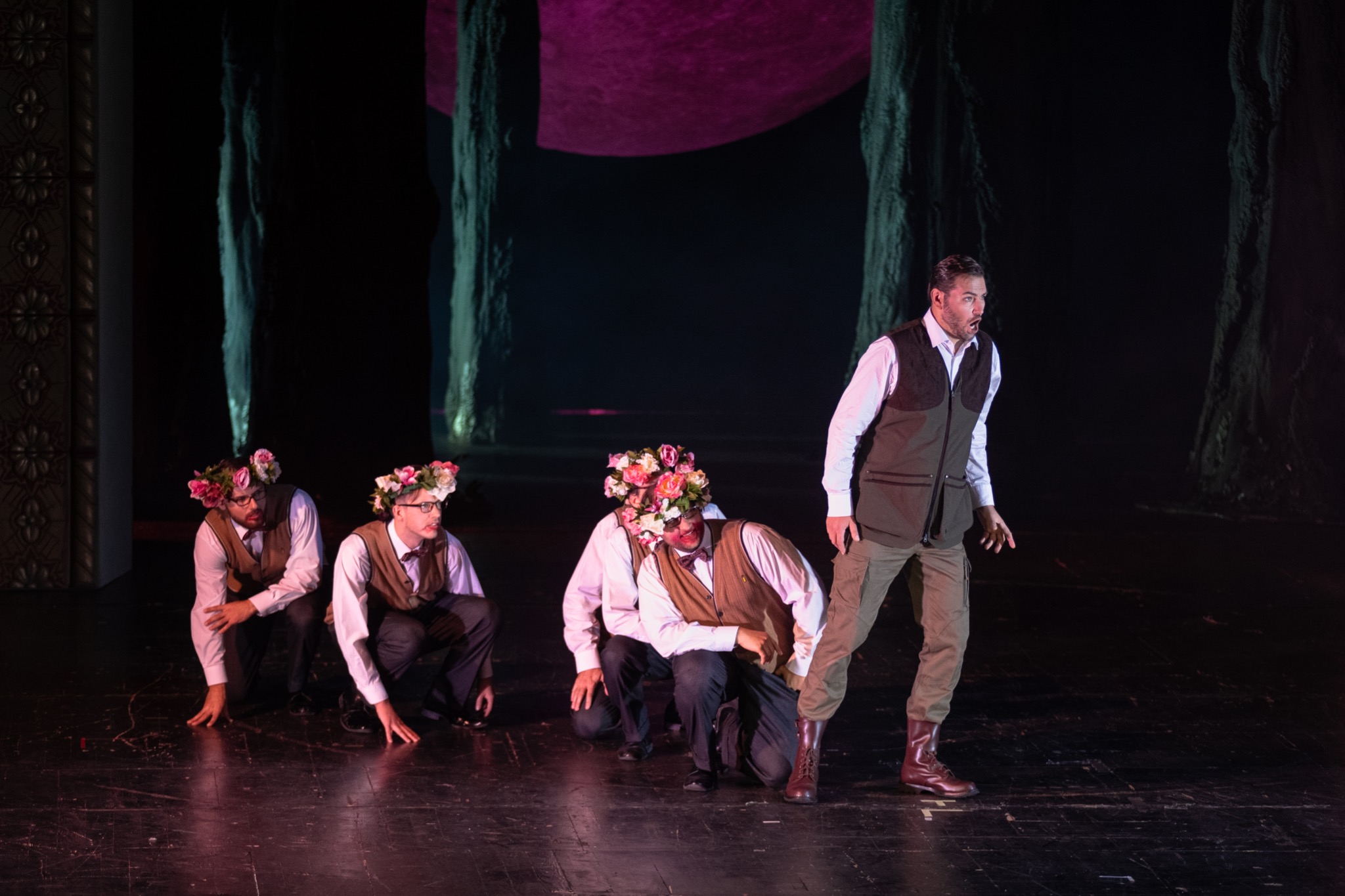 Scena teatralna z czterema aktorami: trzej w pozycji kucznej z wiankami kwiatów na głowach, jeden stojący z wyrazem zaskoczenia, tło imitujące las.