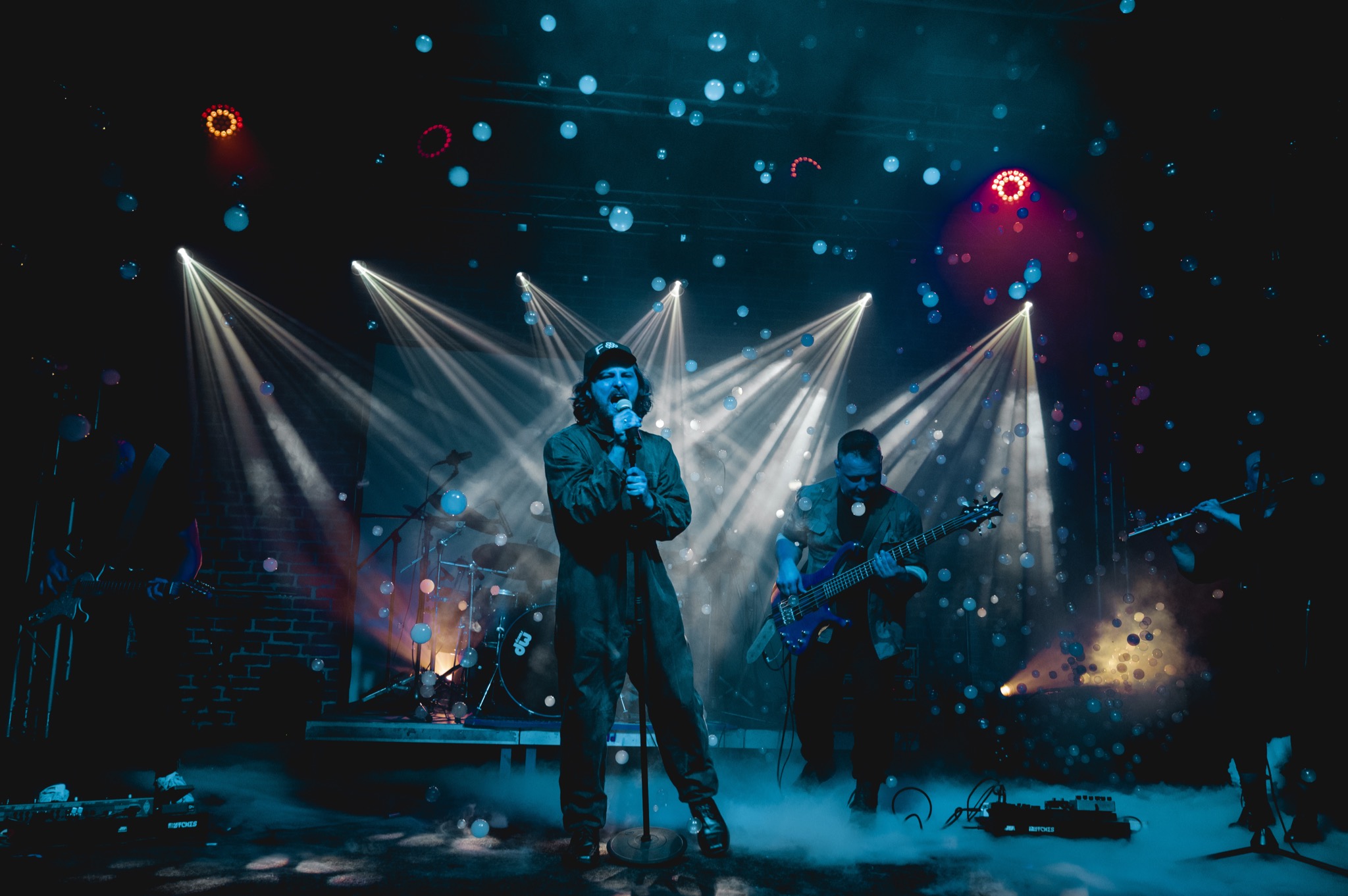 Zespół muzyczny występuje na scenie w oświetleniu scenicznym, z efektem sztucznych baniek mydlanych w powietrzu. Wokalista w kombinezonie i czapce śpiewa do mikrofonu, obok basista gra...