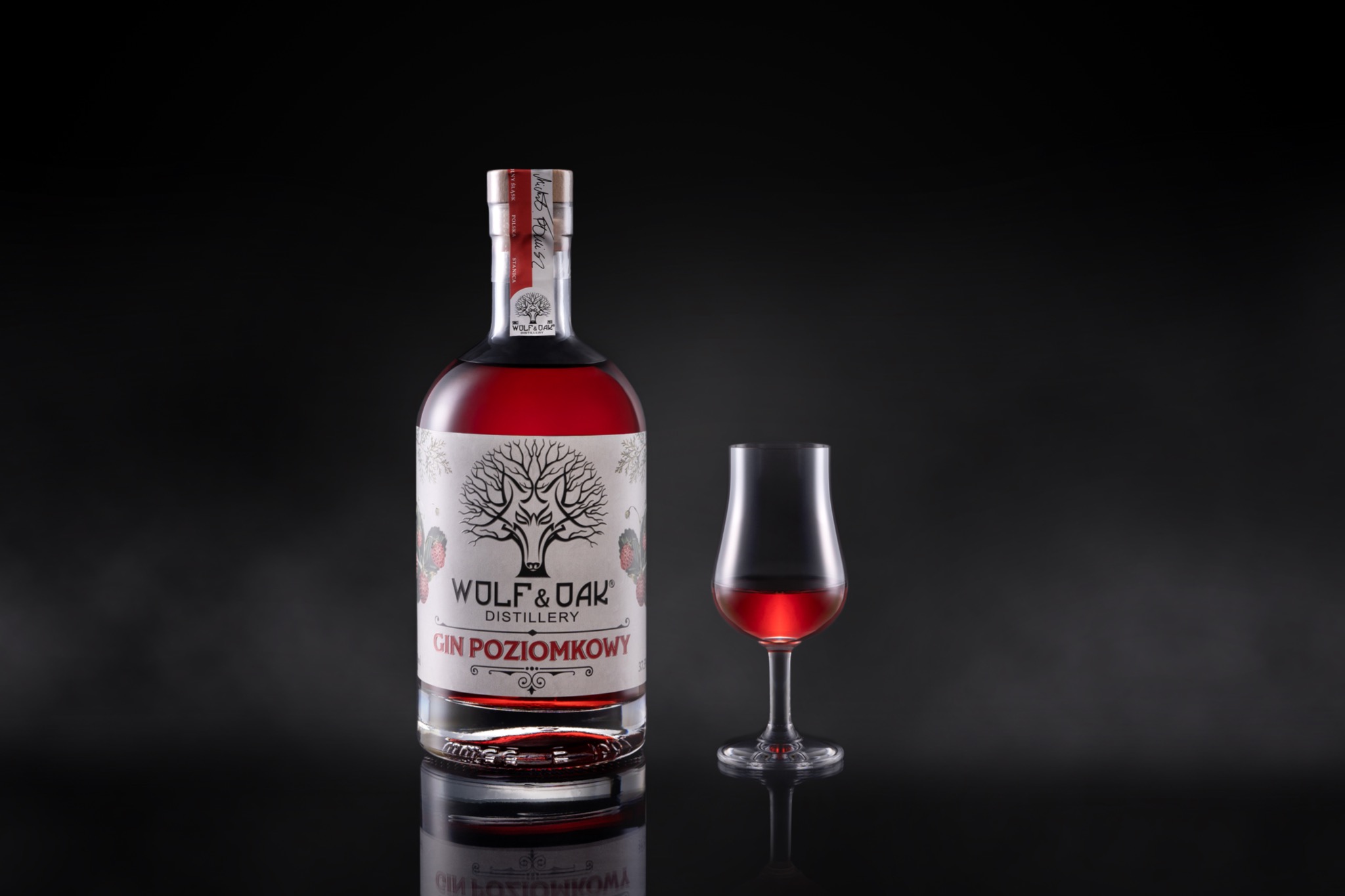 Butelka czerwonego ginu 'Wolf & Oak' z etykietą przedstawiającą drzewo i wilka, obok kieliszek wypełniony tym samym trunkiem, na ciemnym, odbijającym światło tle.