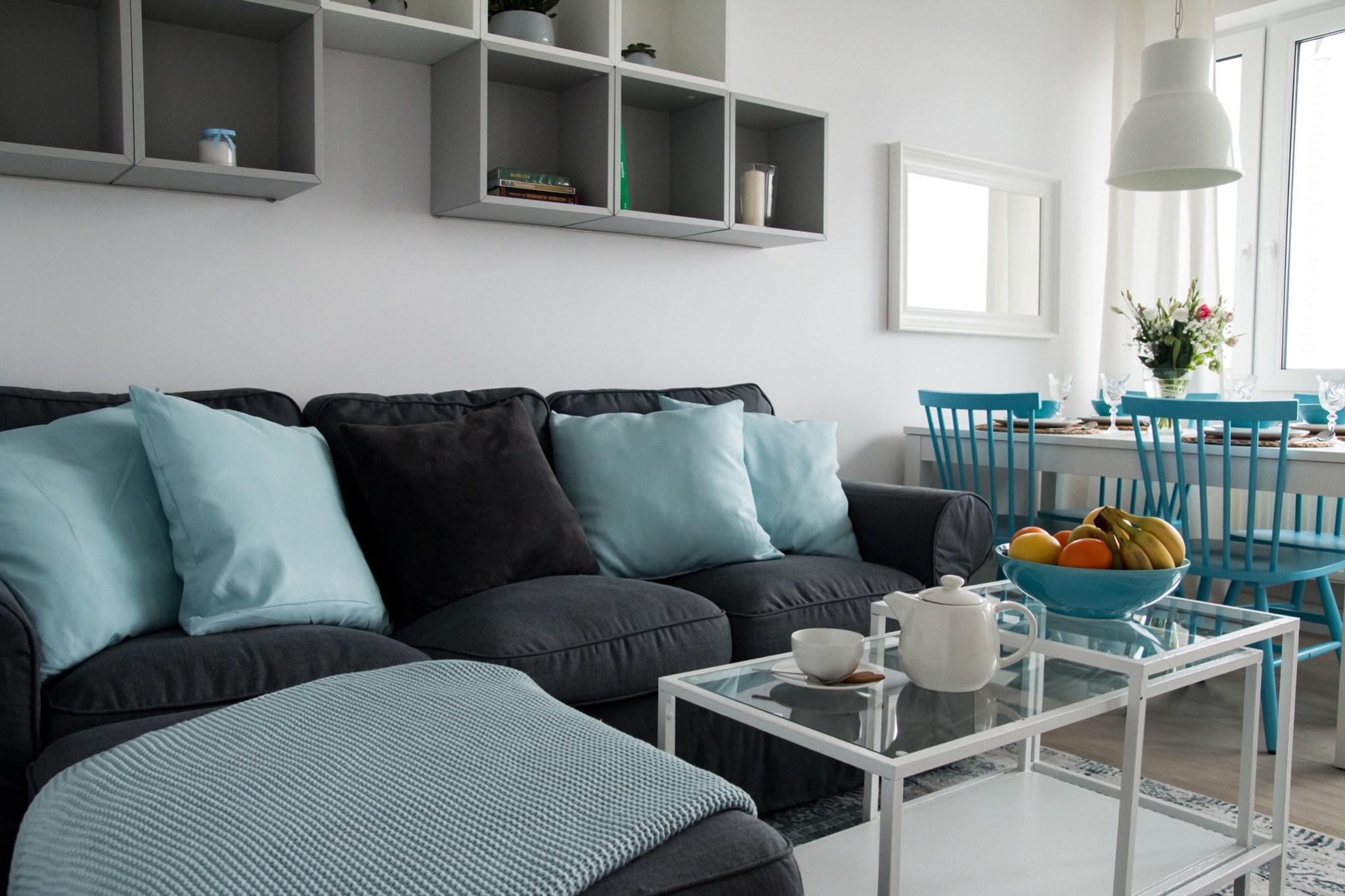 Jasne wnętrze salonu z szarą sofą z turkusowymi i czarnymi poduszkami, szklanym stolikiem kawowym z serwisem do herbaty, jadalnią w tle z turkusowymi krzesłami i owocami w misie, całość...