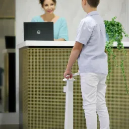 Chłopiec stoi na analizatorze składu ciała, tyłem do kamery, podczas konsultacji, w tle kobieta uśmiecha się znad laptopa.