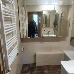 Wyremontowana łazienka z brązowymi płytkami podłogowymi, beżowymi ścianami i białym grzejnikiem drabinkowym, widoczne odbicie osoby robiącej zdjęcie w lustrze.