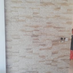 Patryk-Bud - Ściana wykończona dekoracyjną okładziną z jasnego kamienia imitującego piaskowiec, z widocznymi puszkami instalacyjnymi i gniazdami elektrycznymi w trakcie montażu.