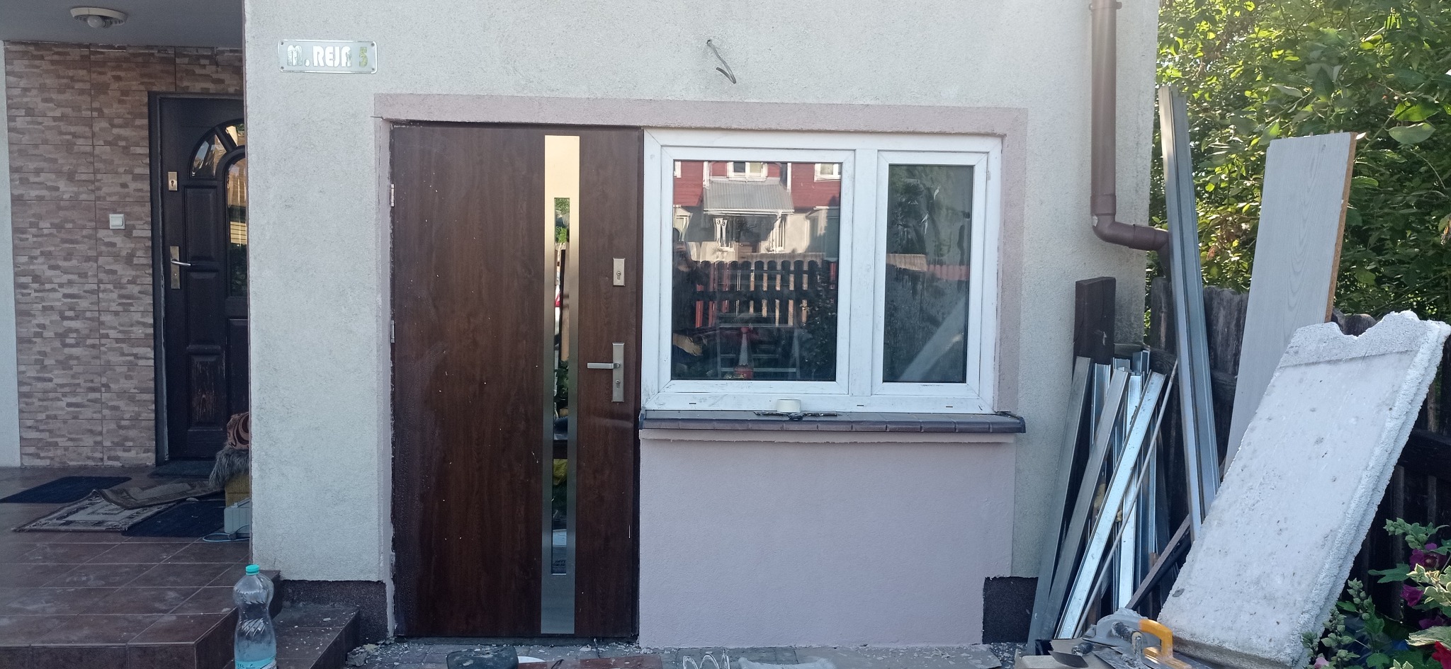 Nowe brązowe drzwi wejściowe z pionowym paskiem stali nierdzewnej i białe okno PCV z widocznym odbiciem budynku, obok leżą materiały budowlane i narzędzia.