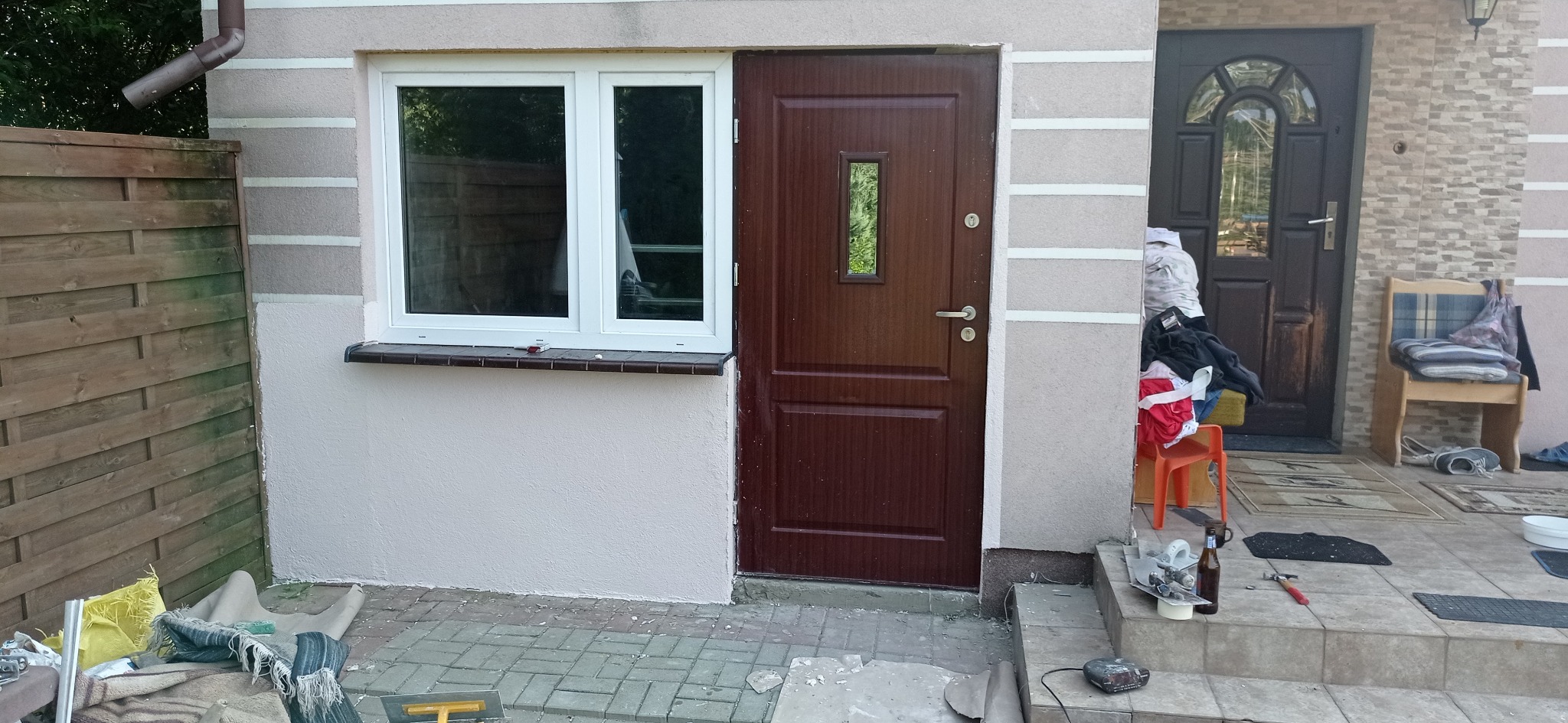 Brązowe drzwi wejściowe z małym okienkiem, białe okno PCV obok, elewacja po nałożeniu tynku strukturalnego, widoczne narzędzia i materiały budowlane na zewnątrz, bałagan przed wejściem.