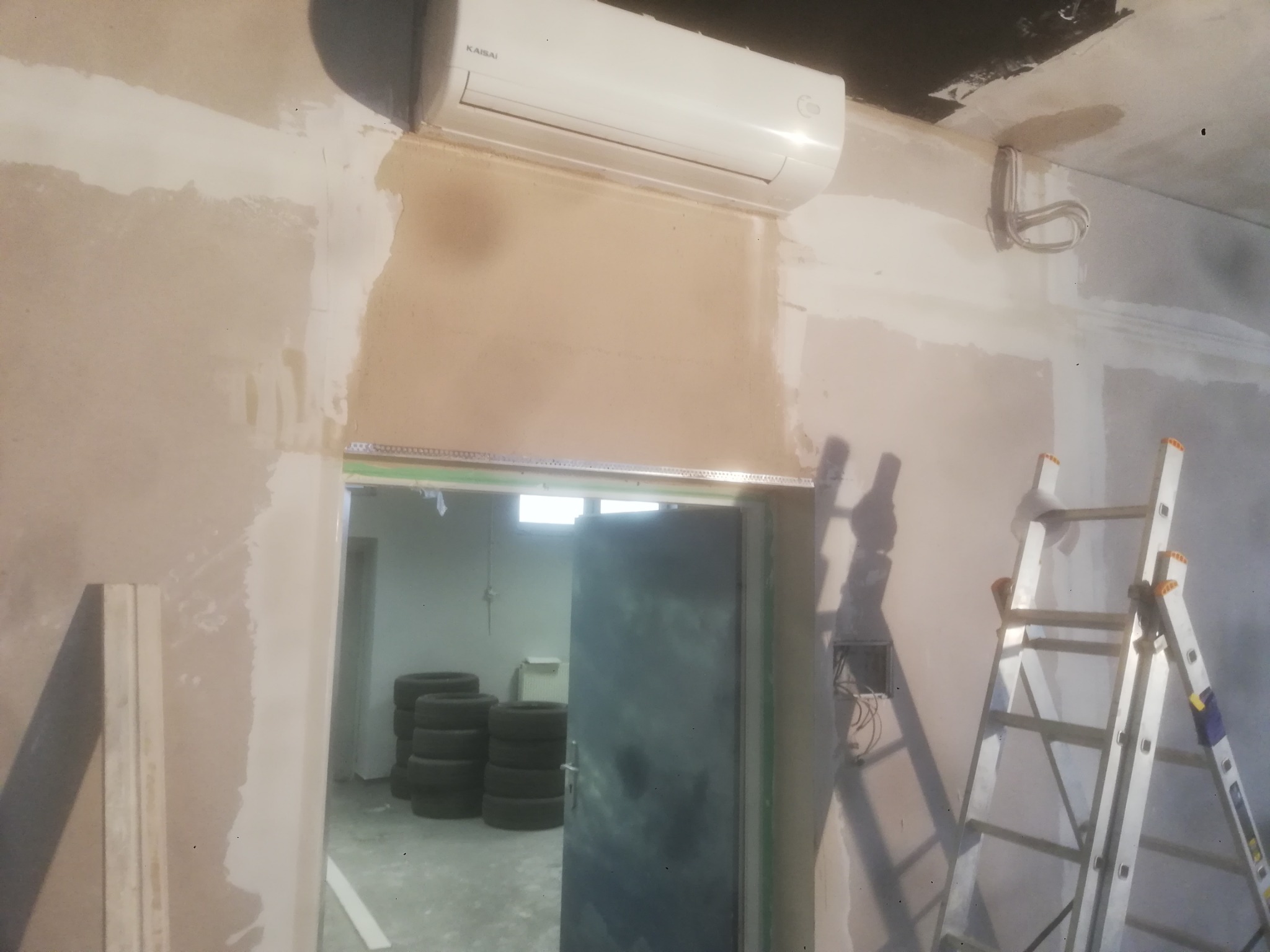 Wnętrze w trakcie prac wykończeniowych: widoczny klimatyzator Kaisai, ściany przygotowane pod gładzie, drabina aluminiowa oraz drzwi do pomieszczenia z oponami.