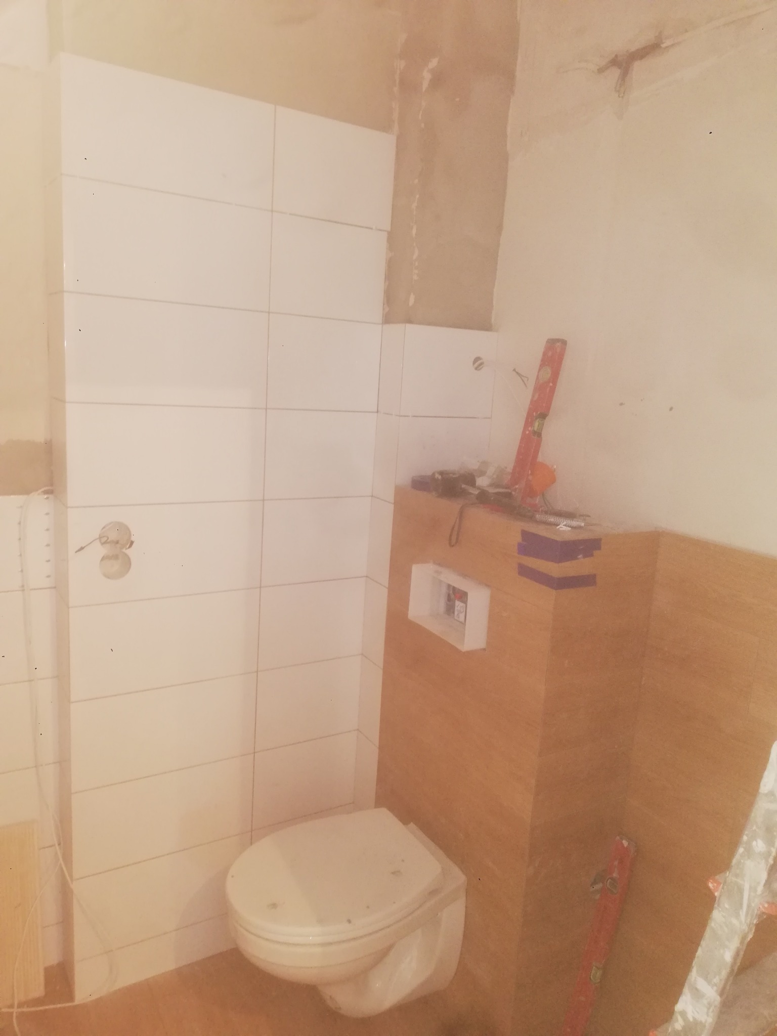 Niedokończona łazienka w trakcie remontu: białe płytki na ścianach, zabudowa WC z imitacją drewna, narzędzia na górze, poziomica oparta o ścianę, widoczne niedociągnięcia w wykończeniu.