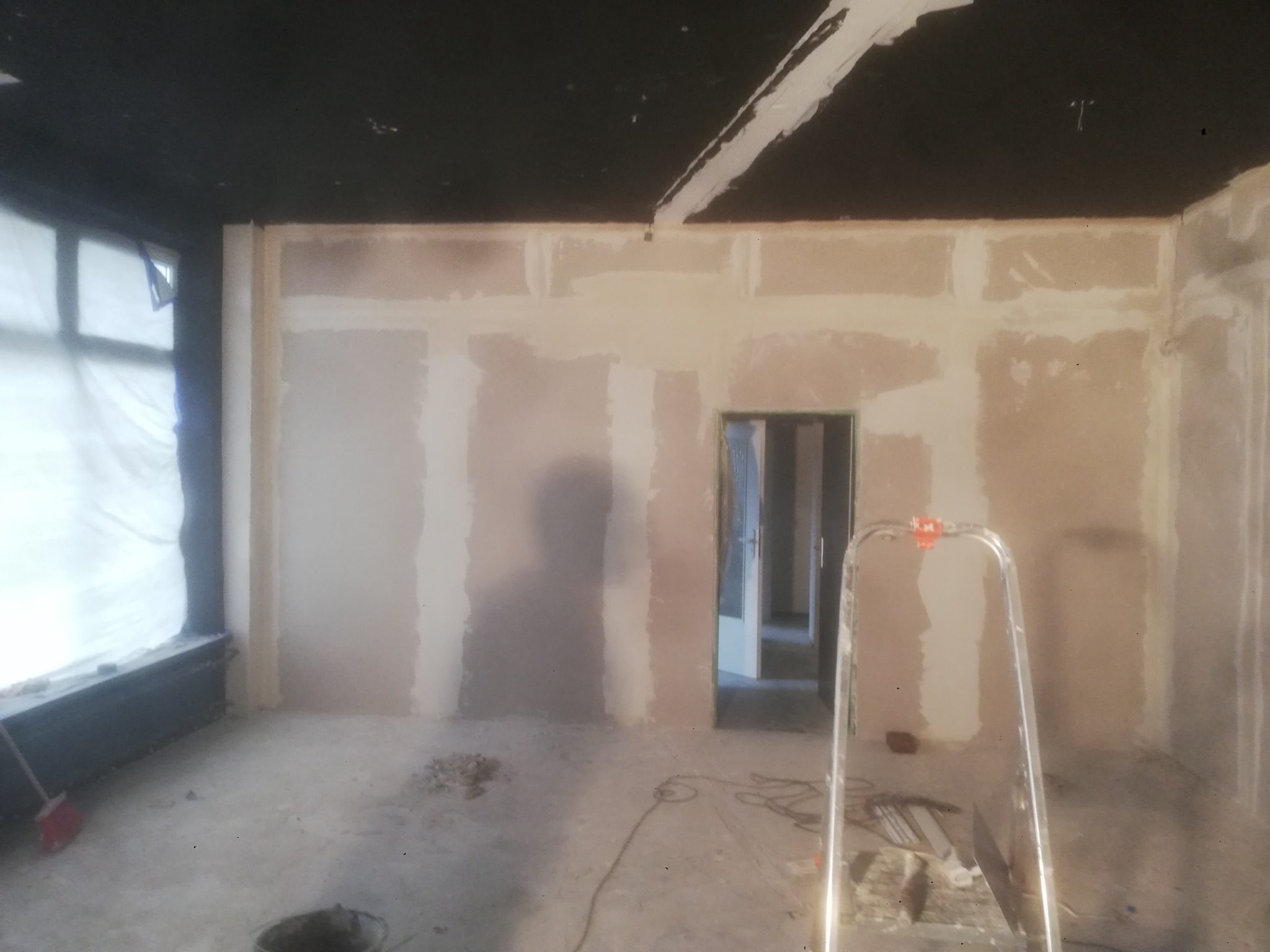 Wnętrze w trakcie prac przygotowawczych do wykończenia: zagruntowane ściany z widocznymi łączeniami płyt gipsowo-kartonowych, czarny sufit z pojedynczymi punktami światła, drabina malarska i wiadro...