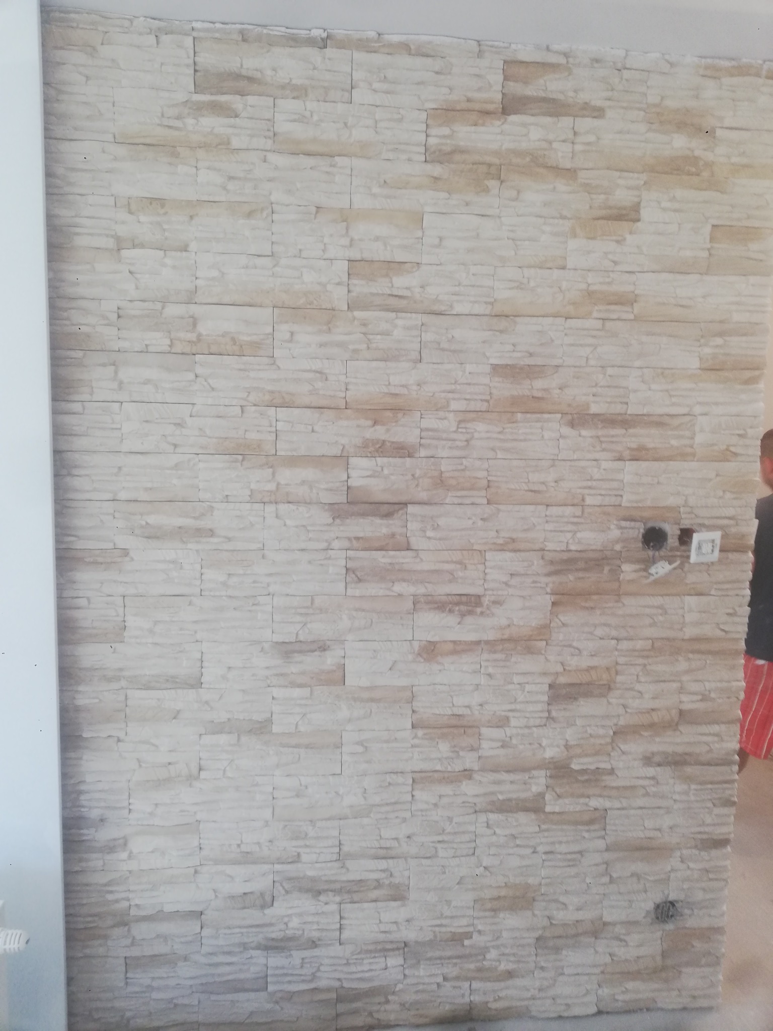 Ściana wykończona dekoracyjną okładziną z jasnego kamienia imitującego piaskowiec, z widocznymi puszkami instalacyjnymi i gniazdami elektrycznymi w trakcie montażu.