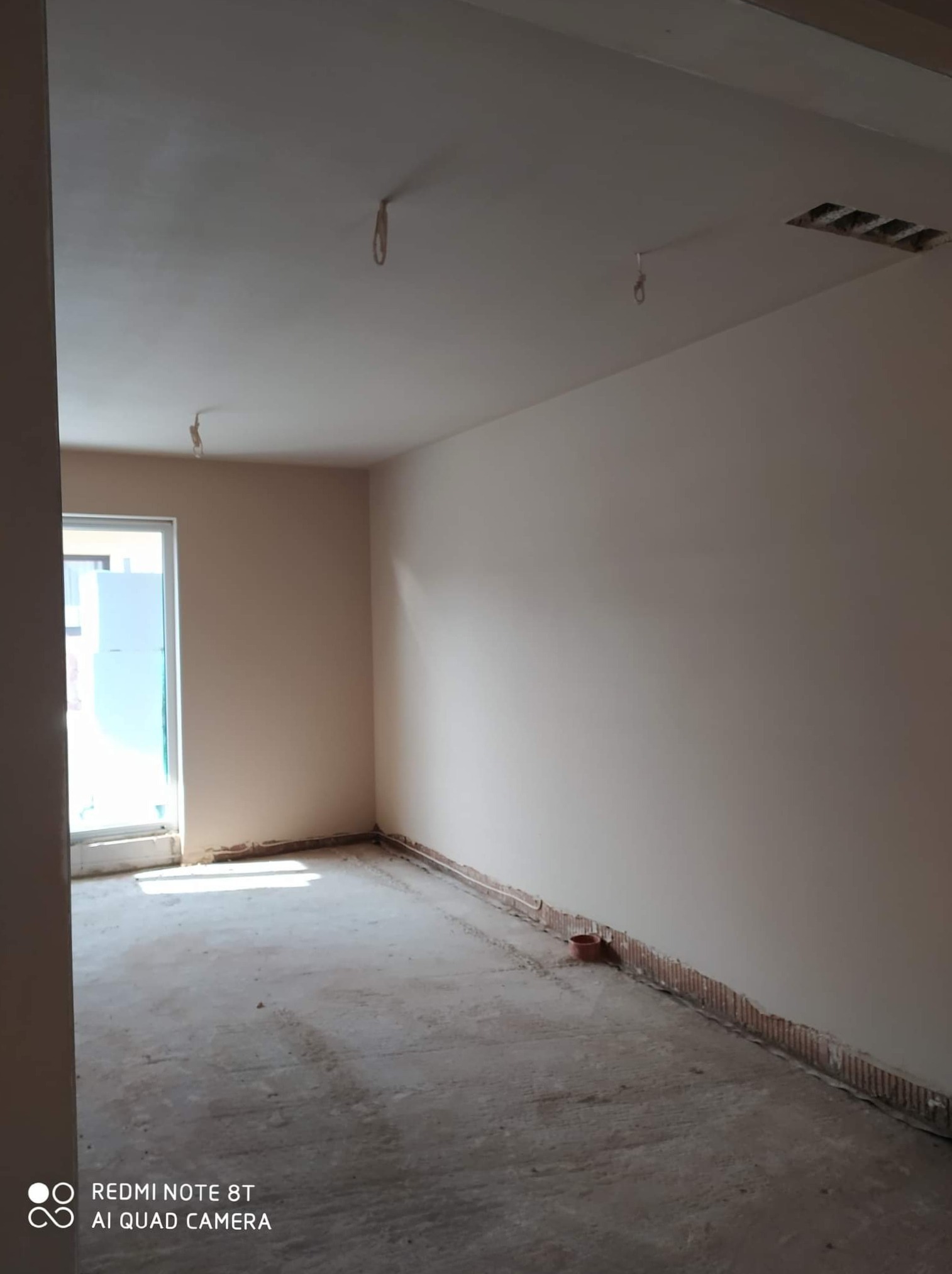 Wykończone ściany w pokoju z widocznymi przewodami wystającymi z sufitu i niedokończoną podłogą, przygotowane do dalszych prac wykończeniowych.