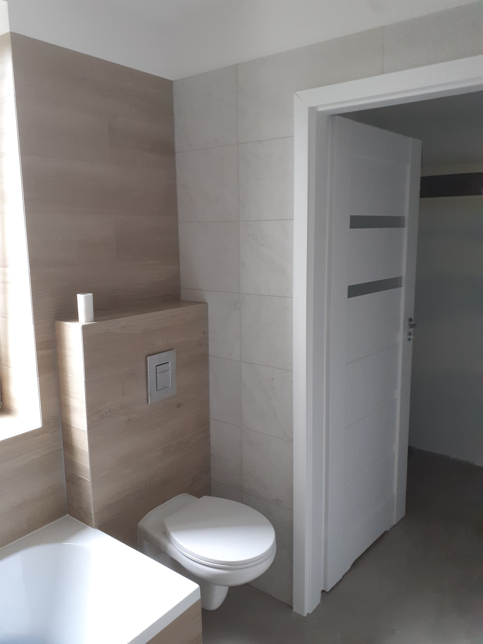 Wykończona łazienka z beżowymi i białymi płytkami, podwieszana toaleta z przyciskiem spłukiwania, biała wanna i białe drzwi z poziomymi wstawkami, widok na niedokończone pomieszczenie przez otwarte...