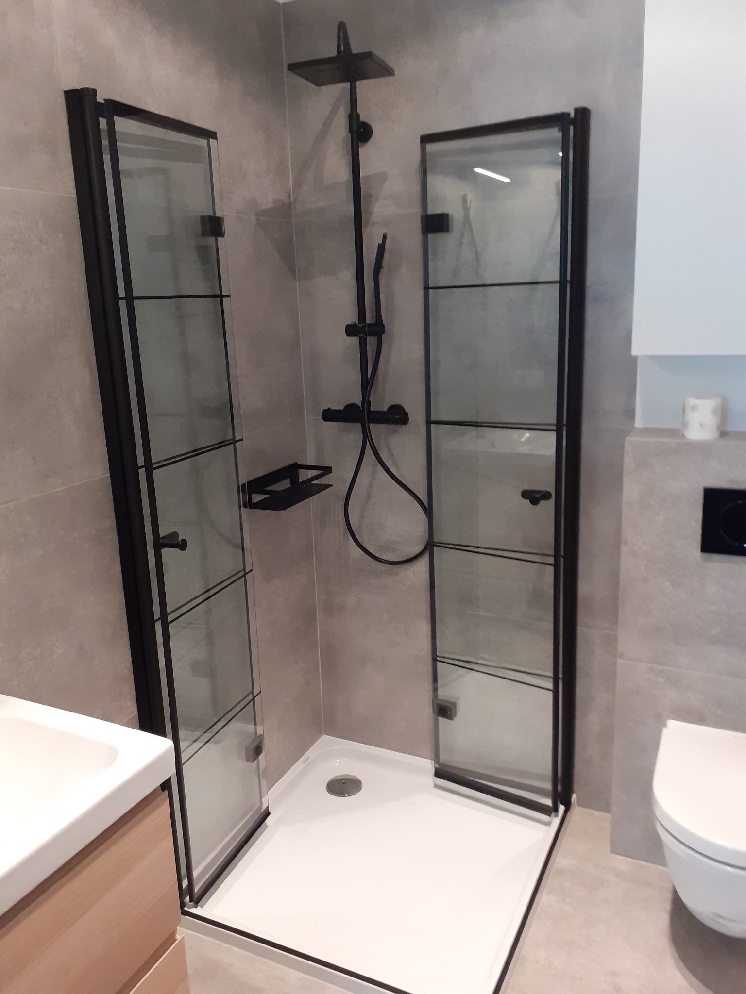 Nowoczesna kabina prysznicowa z czarnymi profilami i szklanymi drzwiami uchylnymi, na białym brodziku, z czarną armaturą, widoczna umywalka z szafką w neutralnych barwach, szara płytka na ścianach.