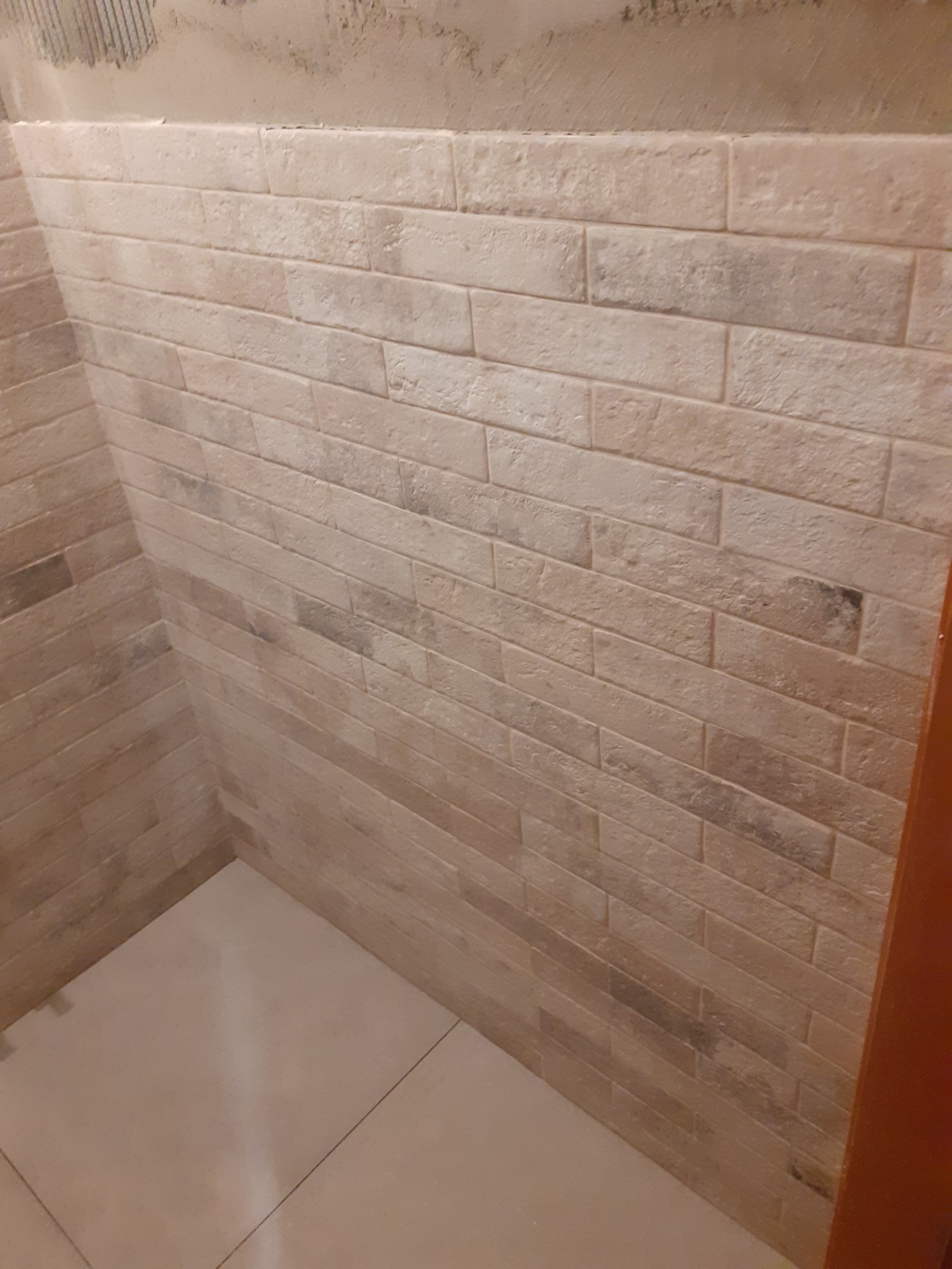 Ściana wyłożona beżowymi płytkami imitującymi cegłę, z widocznymi fugami i fragmentem surowego tynku u góry, oraz jasna, błyszcząca podłoga.