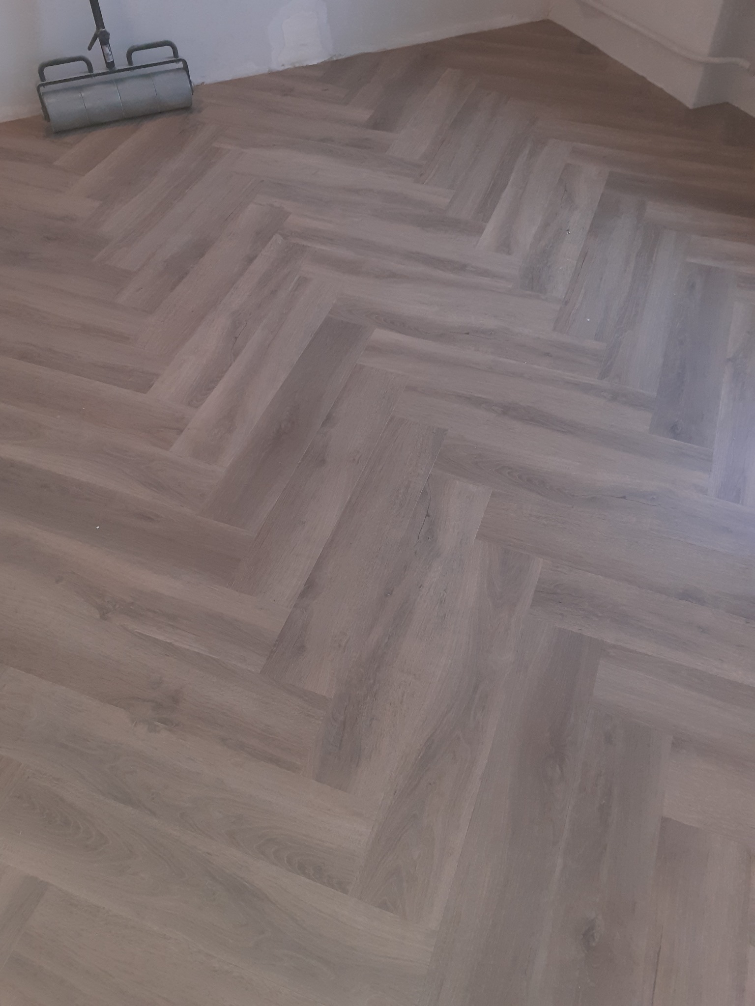 Ułożony parkiet w jodełkę z widocznym wałkiem do malowania przy ścianie.