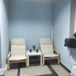 Dwa kremowe fotele z drewnianymi ramami stoją w rogu pokoju o błękitnych ścianach, pomiędzy nimi mały biały stolik z dwoma czarnymi kubkami i małą rośliną doniczkową.