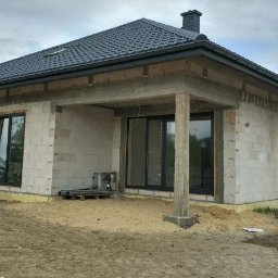 Rafal Zaniewicz - Niedokończony dom jednorodzinny z bloczków, widoczne okna z czarnymi ramami, betonowy strop nad tarasem, czarny dach z kominem, teren wokół domu w trakcie prac ziemnych.