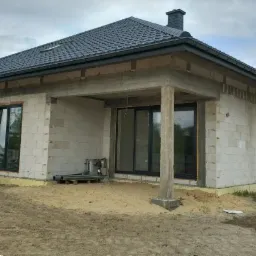 Niedokończony dom jednorodzinny z bloczków, widoczne okna z czarnymi ramami, betonowy strop nad tarasem, czarny dach z kominem, teren wokół domu w trakcie prac ziemnych.