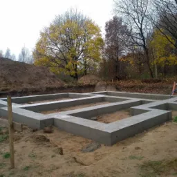 Wylane betonowe fundamenty pod budynek jednorodzinny, widoczne zbrojenia, w tle usypana ziemia i jesienne drzewa.