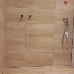 LaSerek Marcin Serafin - Beżowe płytki imitujące drewno na podłodze i ścianach łazienki, widoczne przyłącza wodne i słuchawka prysznicowa, brak fugi w narożniku.