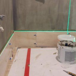 LaSerek Marcin Serafin - Świeżo przygotowana, surowa ściana w łazience z widocznym kranem, wyznaczona laserem linia pozioma, poziomica, wiadro z zaprawą, narzędzia do cięcia płytek i nożyk na podłodze zabezpieczonej folią.