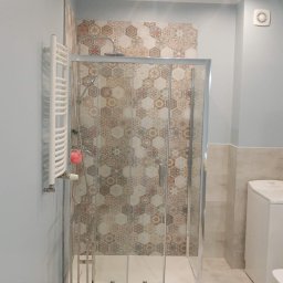 LaSerek Marcin Serafin - Nowoczesna kabina prysznicowa z przesuwnymi drzwiami, wyłożona mozaiką w geometryczne wzory, obok grzejnik drabinkowy z różową gąbką, całość w jasnej łazience z beżowymi płytkami podłogowymi.