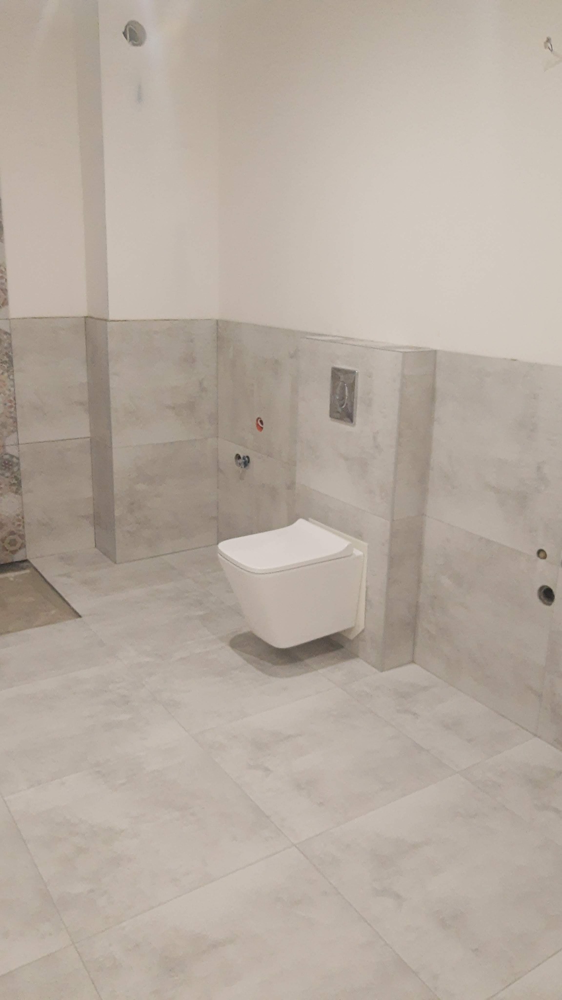 Wisząca miska WC z zabudową w trakcie montażu, widoczne przyłącza wodne i odpływowe, szare płytki na ścianach i podłodze, częściowo ułożone płytki na ścianie po lewej stronie.