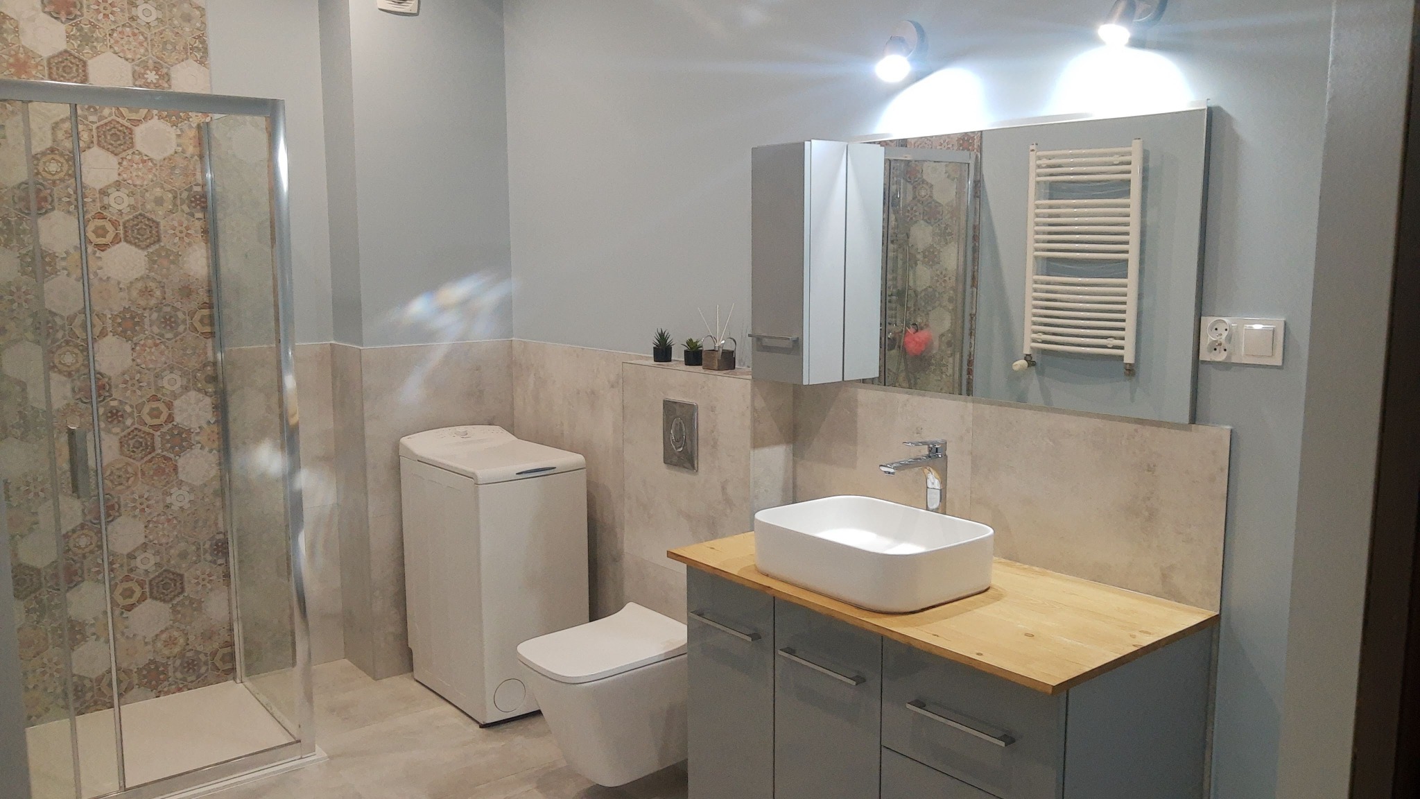 Nowoczesna łazienka w odcieniach szarości i beżu z kabiną prysznicową z heksagonalnymi płytkami, umywalką nablatową na drewnianym blacie i wiszącą toaletą.