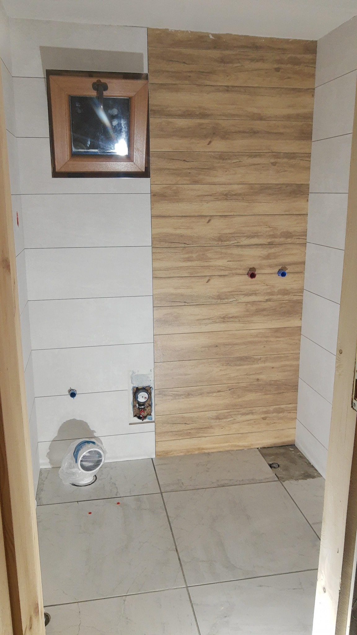 Wnętrze łazienki we Włocławku w trakcie prac wykończeniowych: częściowo ułożone płytki imitujące drewno na ścianie, białe płytki na pozostałej części ściany i podłodze, widoczne instalacje...