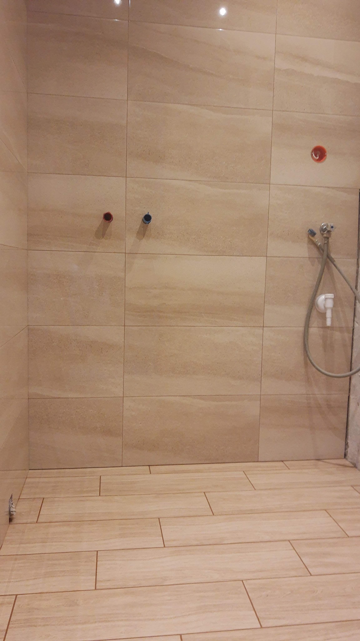 Beżowe płytki imitujące drewno na podłodze i ścianach łazienki, widoczne przyłącza wodne i słuchawka prysznicowa, brak fugi w narożniku.