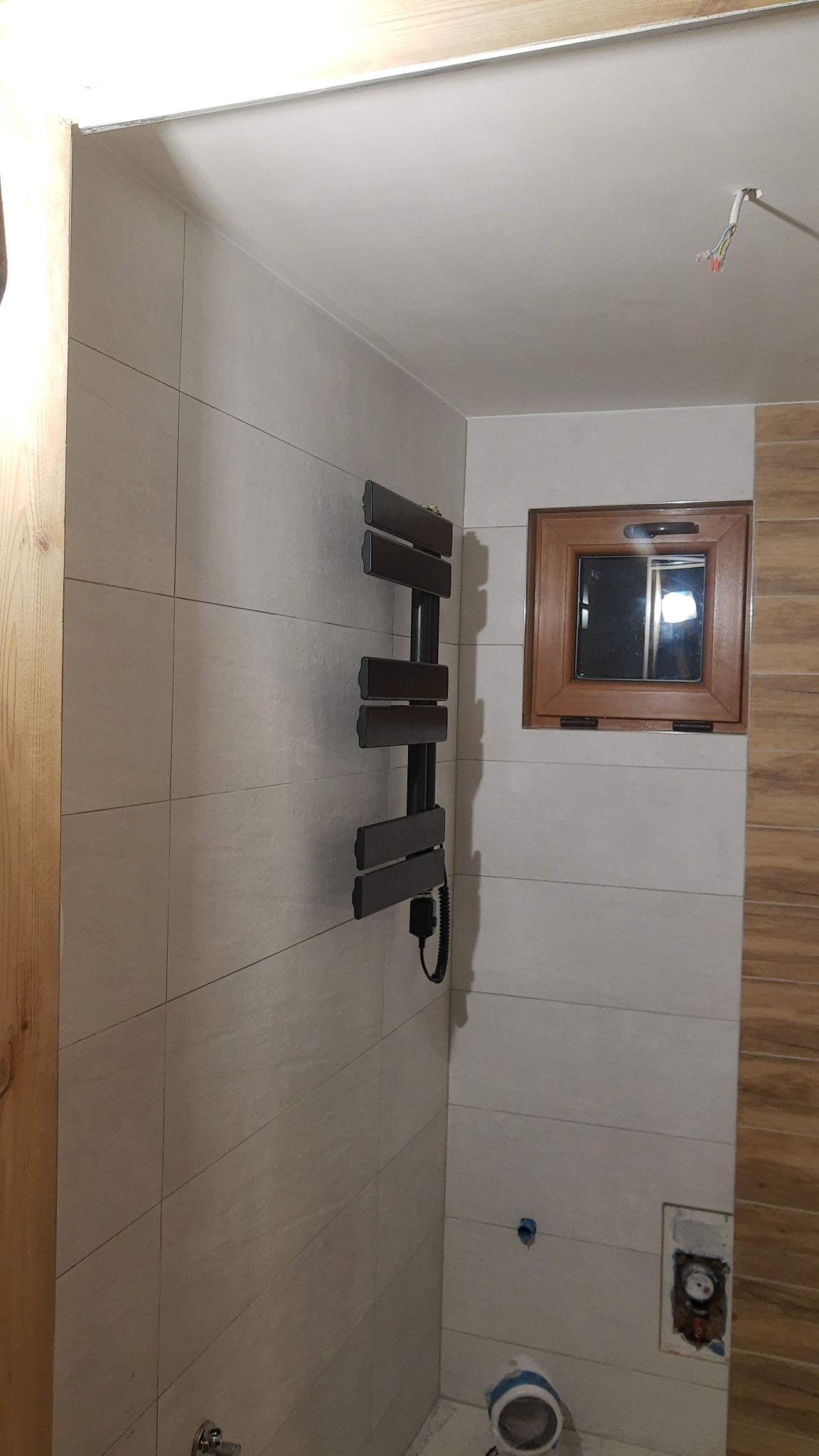 Beżowe płytki ścienne w łazience z zamontowanym, ciemnym grzejnikiem drabinkowym i drewnianym oknem, widoczne niedokończone przyłącza wodno-kanalizacyjne.