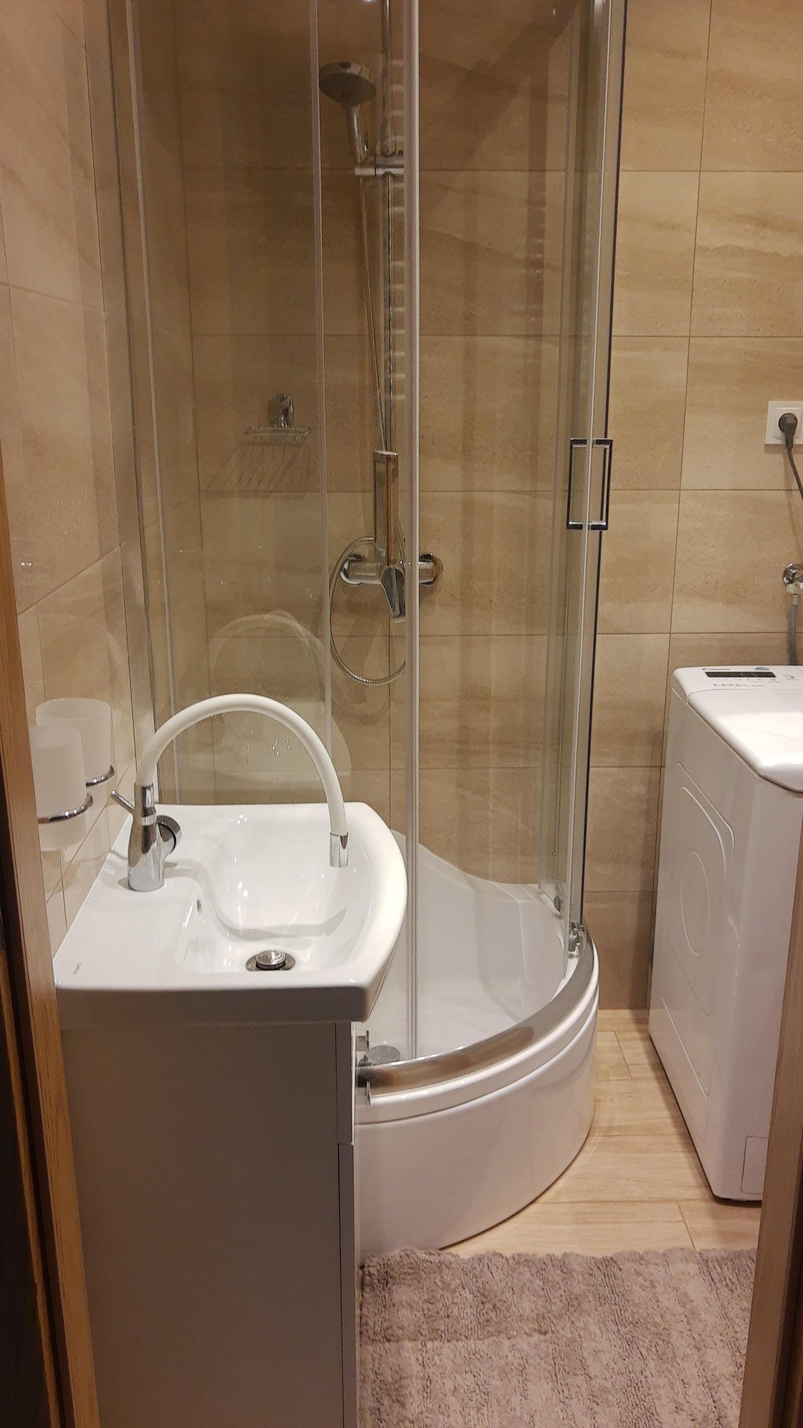 Wnętrze łazienki z kabiną prysznicową narożną, umywalką z szafką i pralką, wykończone beżowymi płytkami i brązowym dywanikiem.