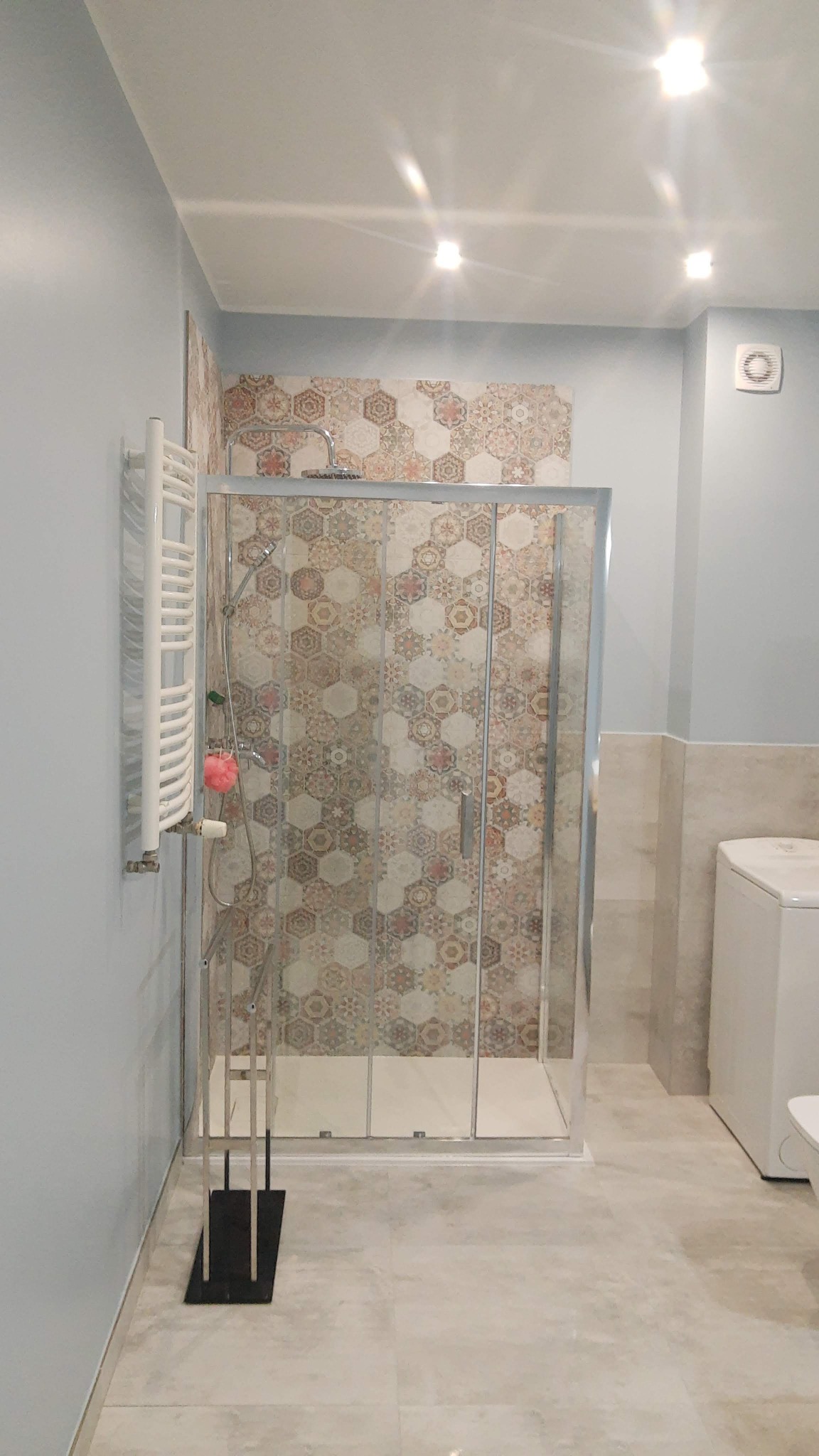 Nowoczesna kabina prysznicowa z przesuwnymi drzwiami, wyłożona mozaiką w geometryczne wzory, obok grzejnik drabinkowy z różową gąbką, całość w jasnej łazience z beżowymi płytkami podłogowymi.