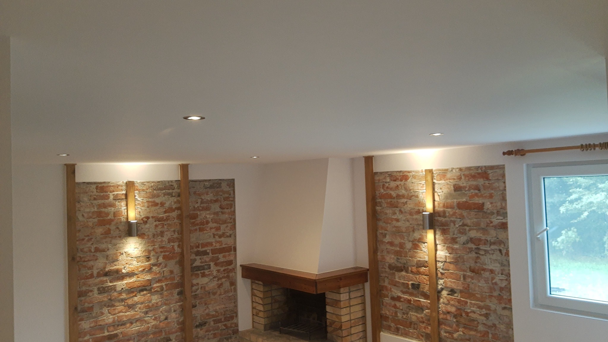 Wnętrze z ceglaną ścianą, drewnianymi elementami i kominkiem we Włocławku, oświetlone punktowymi lampami sufitowymi i kinkietami.
