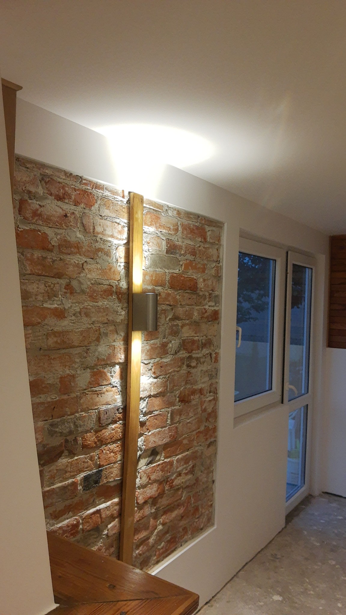 Ściana z czerwonej cegły z zamontowanym pionowym oświetleniem LED na drewnianej listwie, widok na okno i fragment drewnianego blatu.