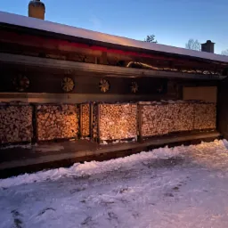 Zadaszony skład z ułożonymi rzędami pociętego drewna opałowego, widoczne wentylatory w suficie, śnieg na ziemi i o zmierzchu.