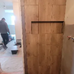 Ściana wykończona panelami imitującymi drewno z wnęką, widok na remontowane pomieszczenie z pracownikiem w tle, podłoga zabezpieczona taśmą malarską, pędzel malarski na podłodze.