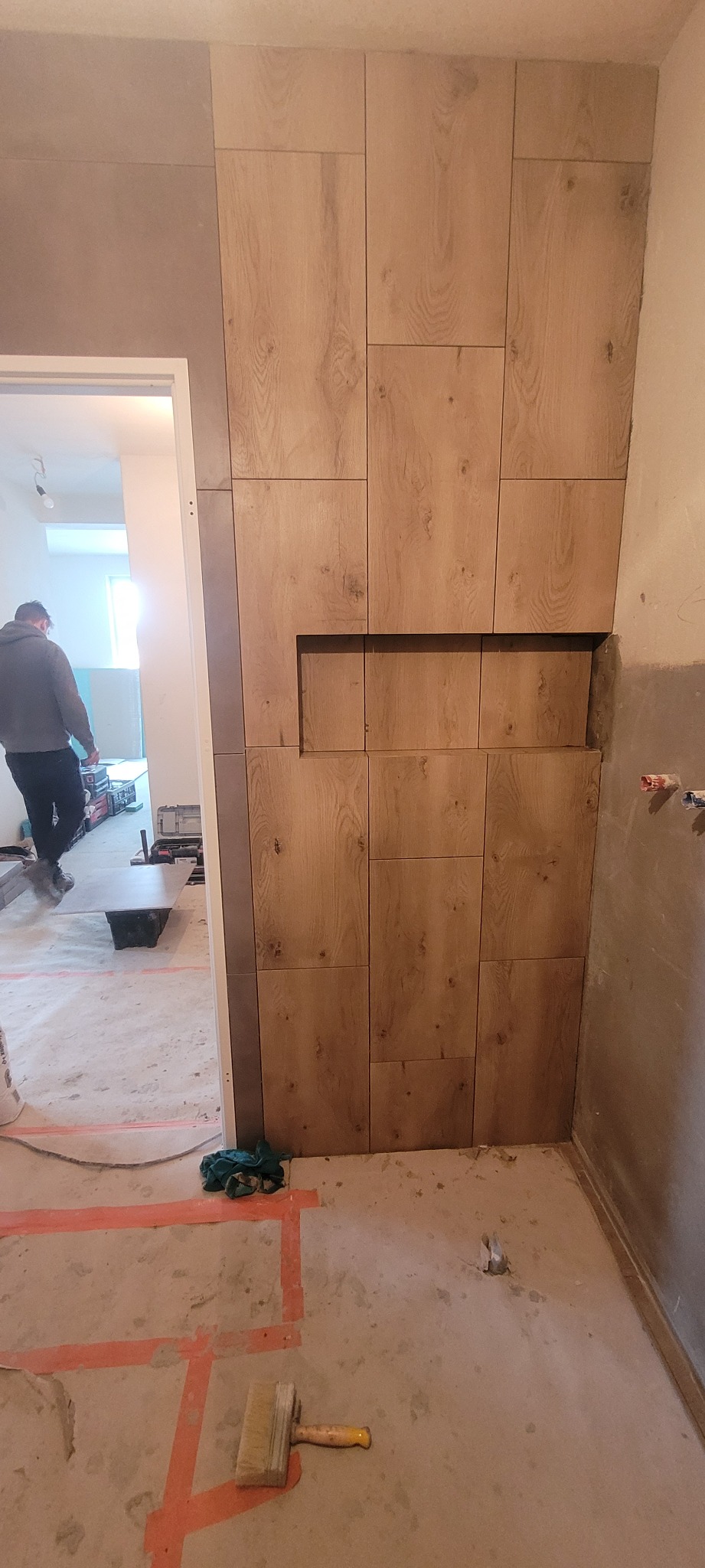 Ściana wykończona panelami imitującymi drewno z wnęką, widok na remontowane pomieszczenie z pracownikiem w tle, podłoga zabezpieczona taśmą malarską, pędzel malarski na podłodze.