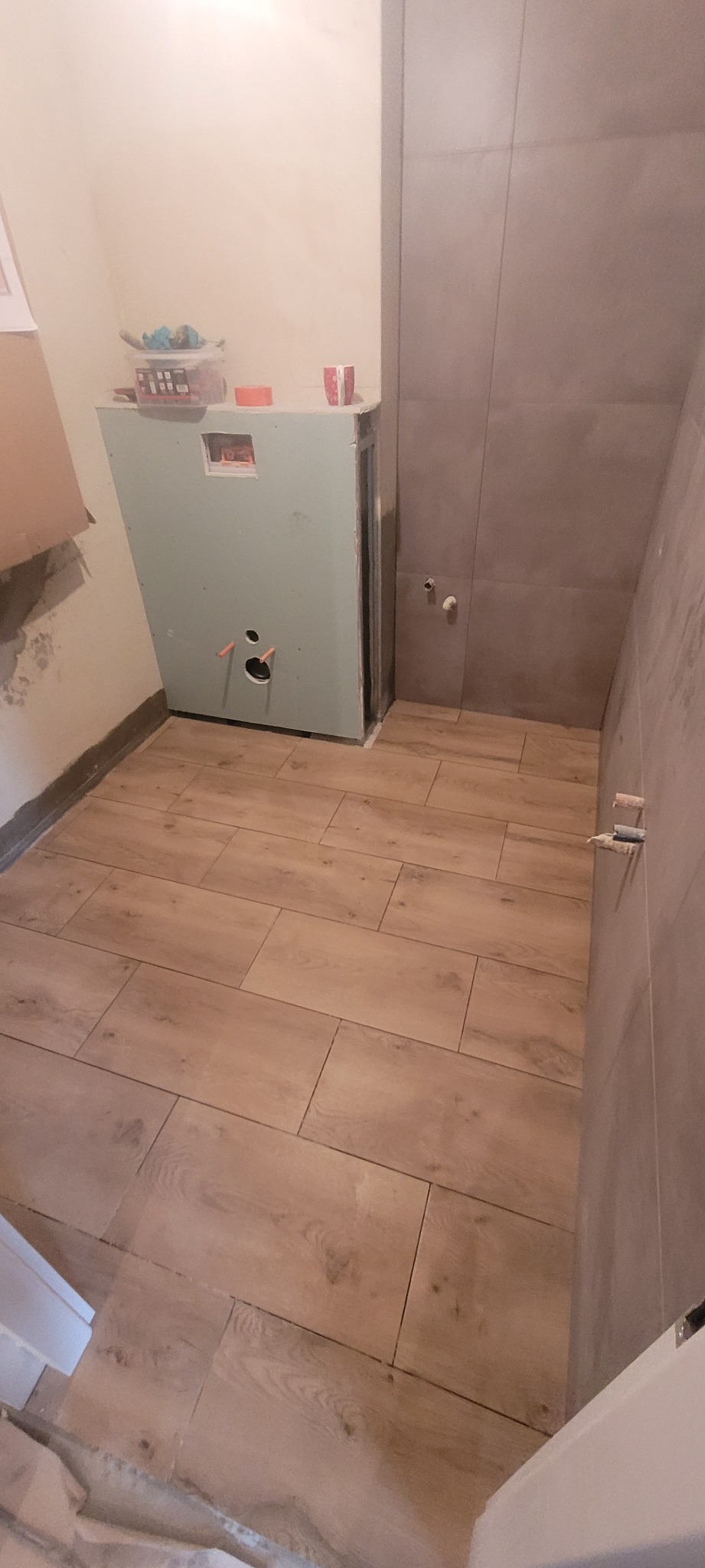 Wnętrze łazienki w trakcie prac: zabudowa stelaża WC z płyty gipsowo-kartonowej, częściowo wykończona ściana z szarych płytek, podłoga wyłożona panelami imitującymi drewno.