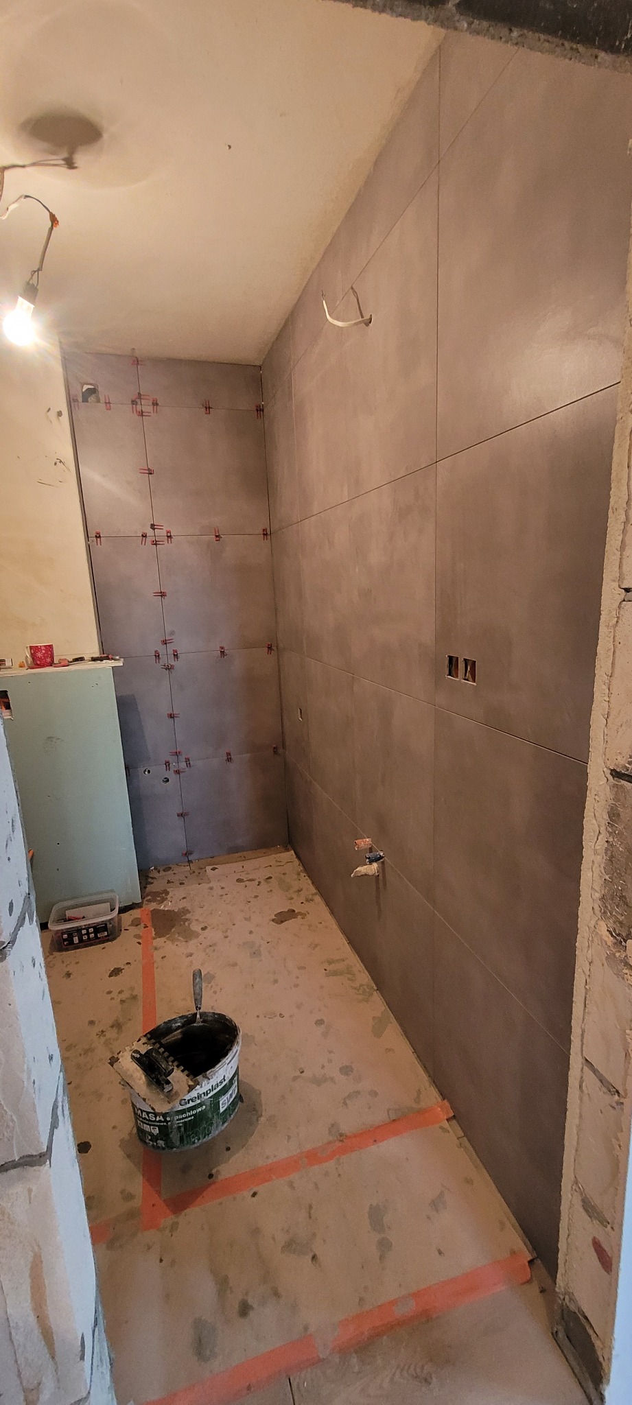 Łazienka w trakcie kładzenia szarych płytek, widoczne krzyżyki dystansowe, wiadro z klejem na podłodze i wyznaczone linie układania płytek.