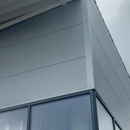 Sg-Bud - Fragment ściany hali przemysłowej wykończonej szarymi płytami warstwowymi, widoczne okna w czarnych ramach i fragment dachu