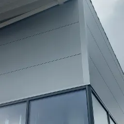 Fragment ściany hali przemysłowej wykończonej szarymi płytami warstwowymi, widoczne okna w czarnych ramach i fragment dachu