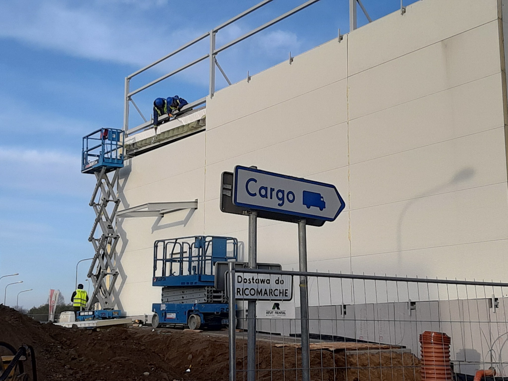 Montaż elewacji z płyt warstwowych na hali w Międzyrzecu Podlaskim, widoczni pracownicy na podnośniku koszowym, obok znak informacyjny 'Cargo' i 'Dostawa do Ricomarche'.