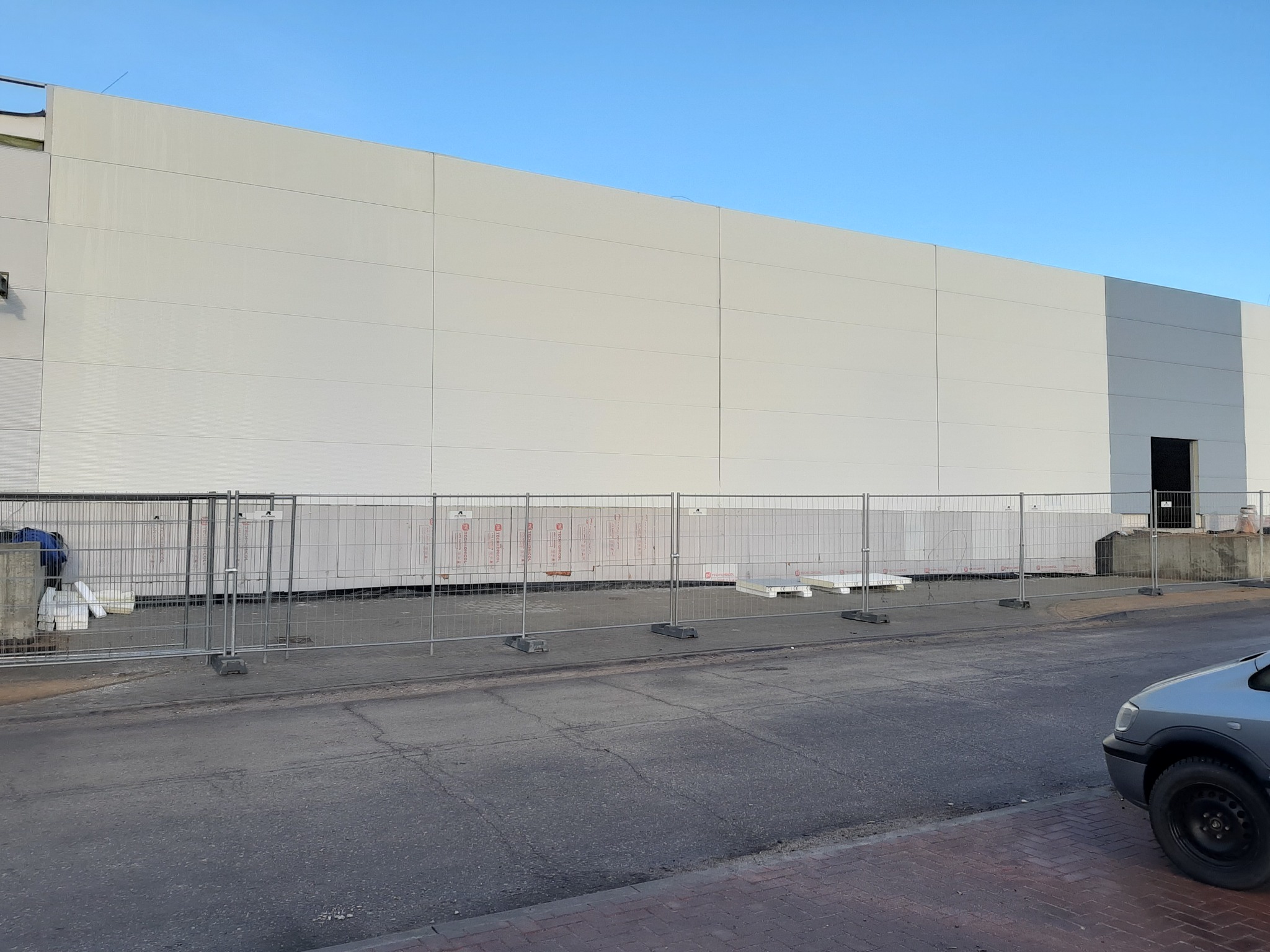 Montaż elewacji hali z jasnych płyt warstwowych w trakcie budowy, widoczne zabezpieczenia budowlane i fragment zaparkowanego auta z przodu