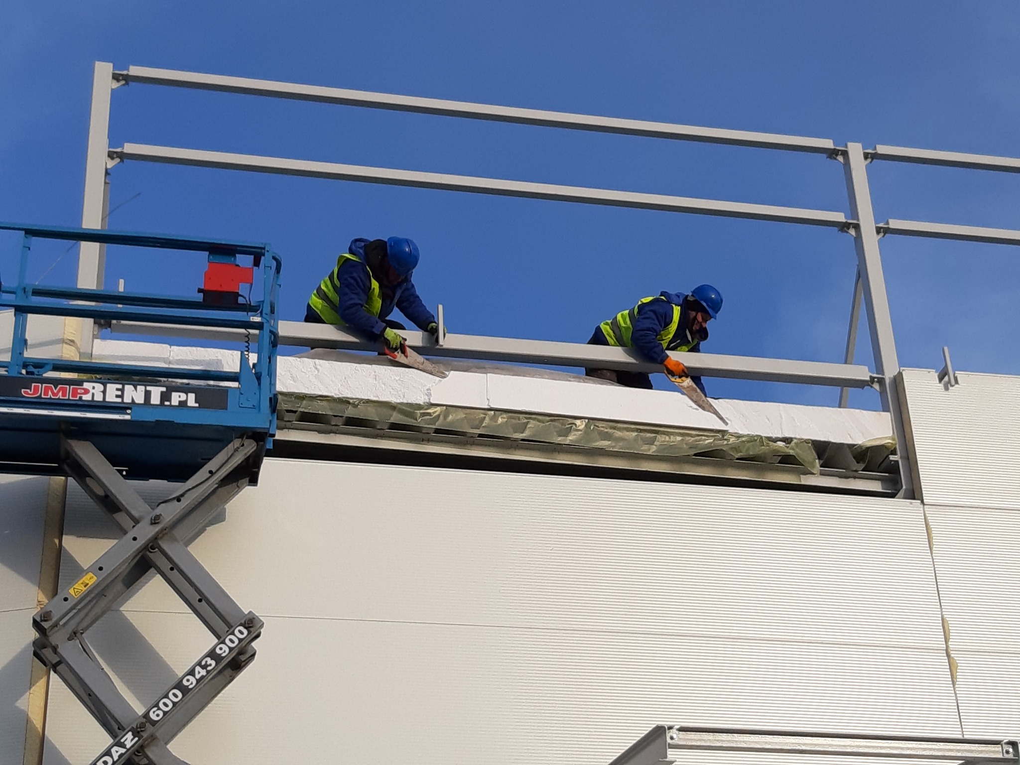 Montaż płyt warstwowych na elewacji hali; dwóch pracowników w kaskach i kamizelkach na podnośniku tnie piłą ręczną białe płyty izolacyjne, widoczny fragment elewacji z płyt warstwowych oraz stalowa...