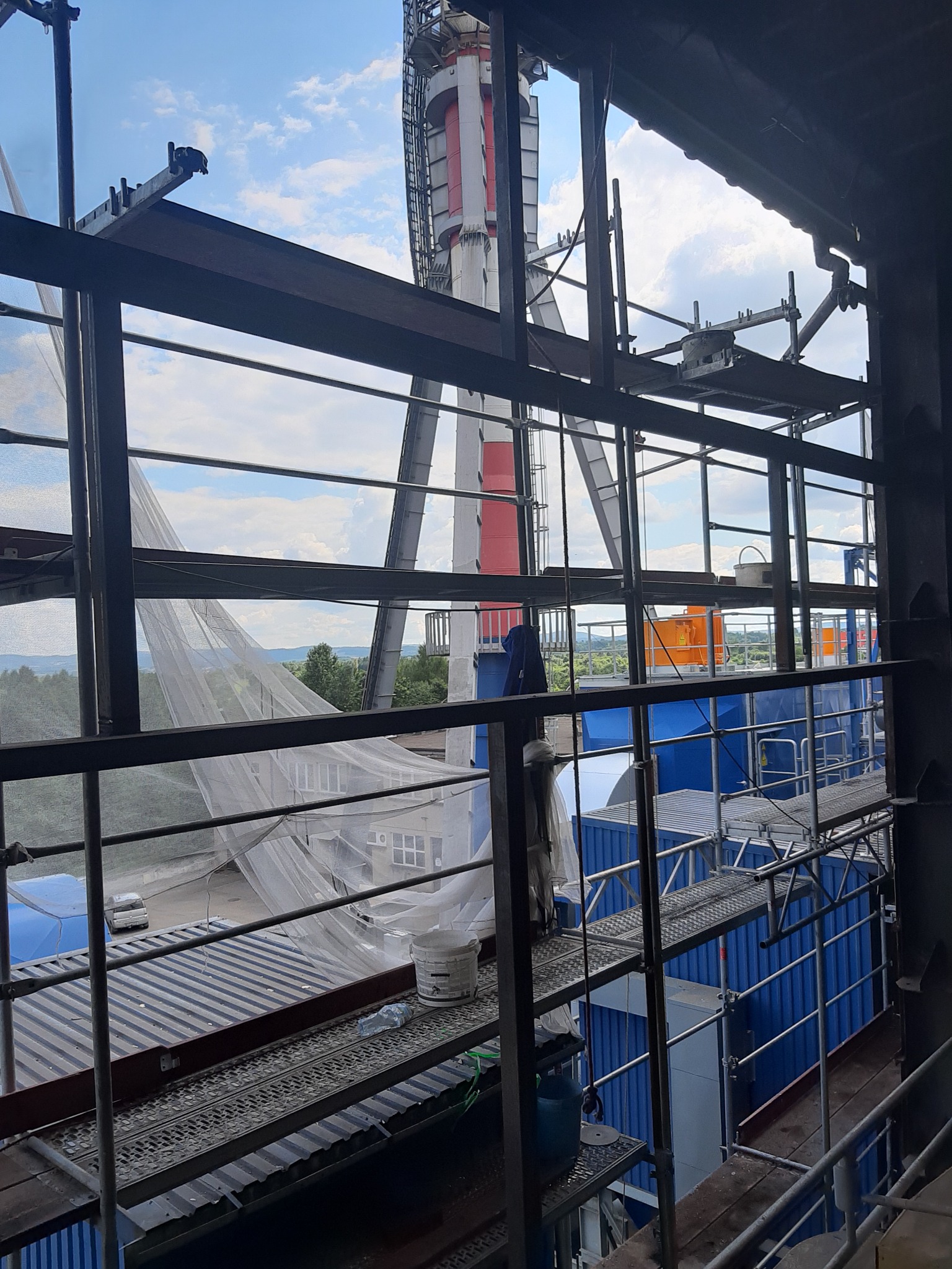 Wnętrze konstrukcji stalowej w trakcie budowy, widoczne rusztowania, elementy niebieskiej hali w tle oraz komin z czerwoną górną częścią.