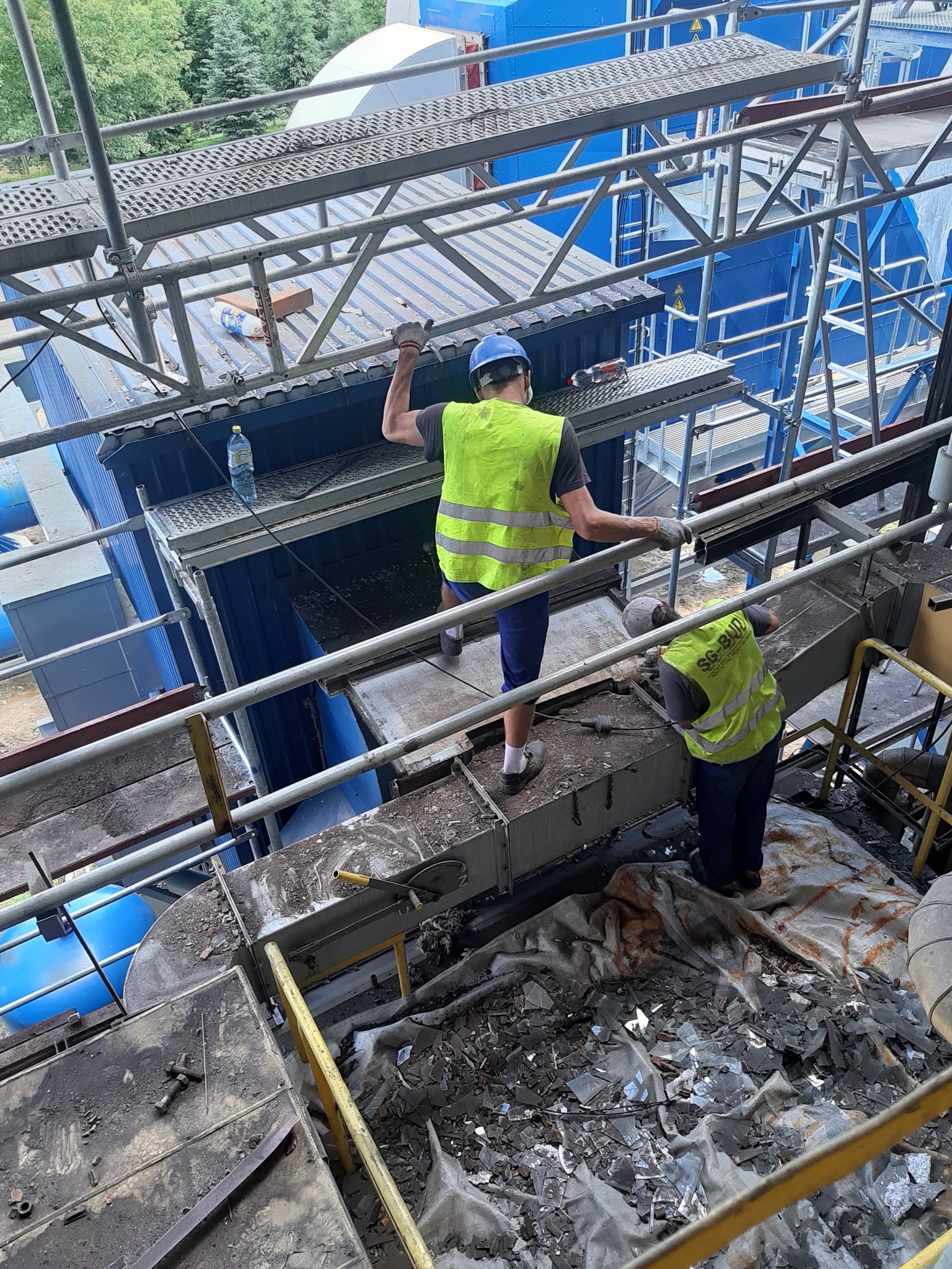 Dwóch pracowników w kamizelkach odblaskowych i kaskach pracuje na wysokościach przy montażu konstrukcji stalowej hali, widoczne rusztowania i elementy konstrukcyjne.