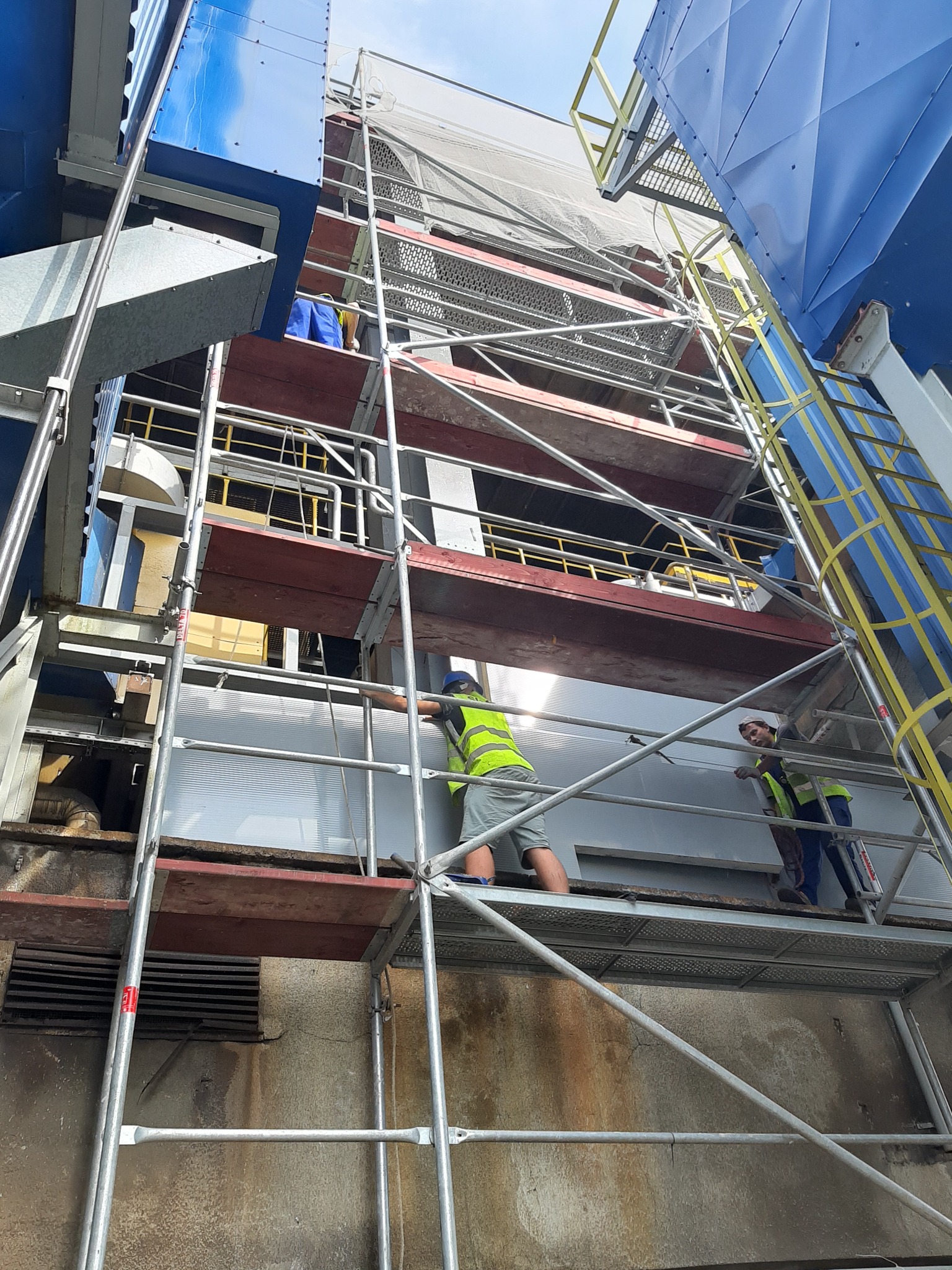 Dwóch pracowników na rusztowaniu przy wysokiej konstrukcji stalowej, prawdopodobnie podczas montażu lub konserwacji, widok z dołu.